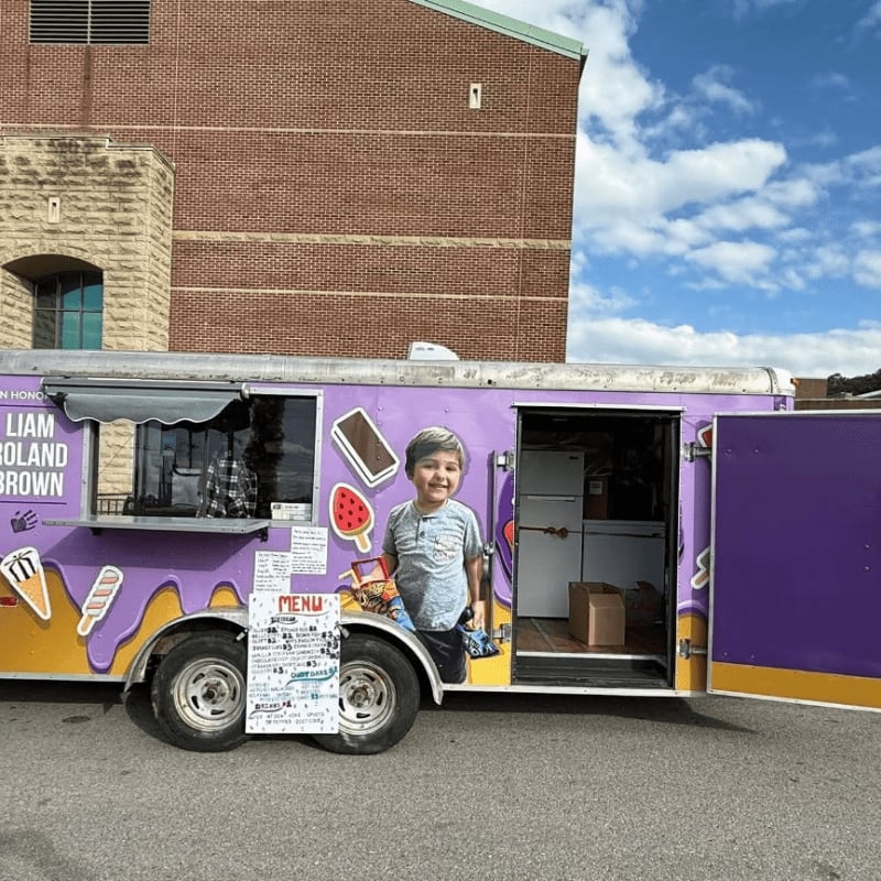 A young boy stands in the doorway of a purple ice cream truck parked in front of a large brick building under a blue sky.