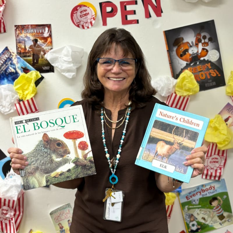 A smiling woman holds two nature books in the foreground, standing before a wall decorated with other books and colorful ornaments.