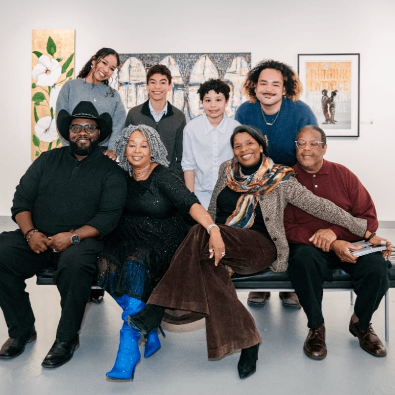 A group of seven people pose together in a brightly lit room featuring artwork on the white walls.