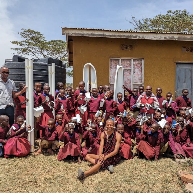 A group of students in maroon uniforms and a few adults pose with white pipes in front of a yellow building and a large black water tank.