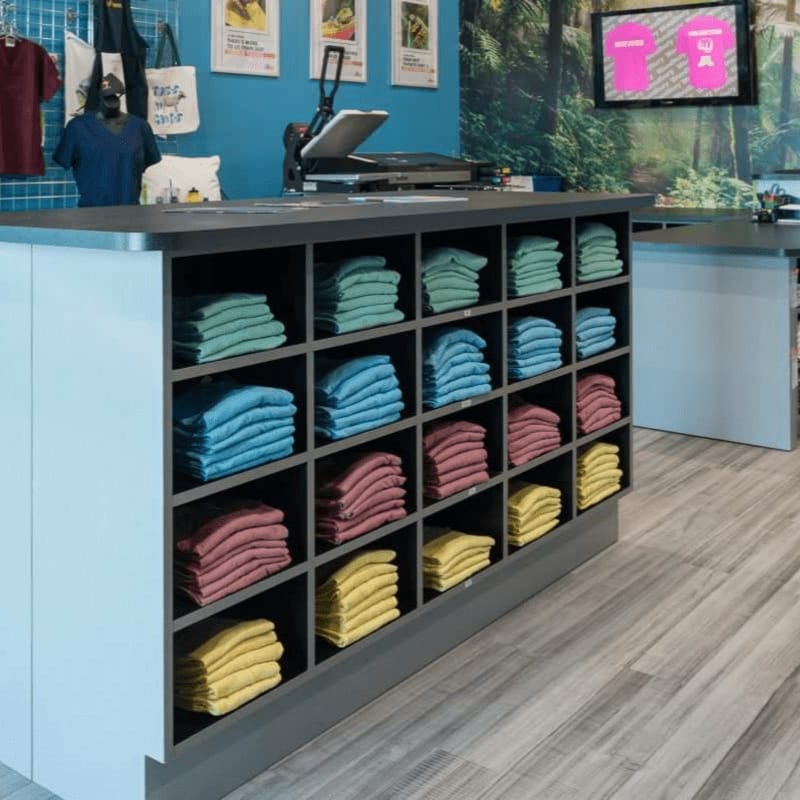 A grey shelving unit filled with neatly folded colorful t-shirts stands in the foreground of a blue-walled clothing store.
