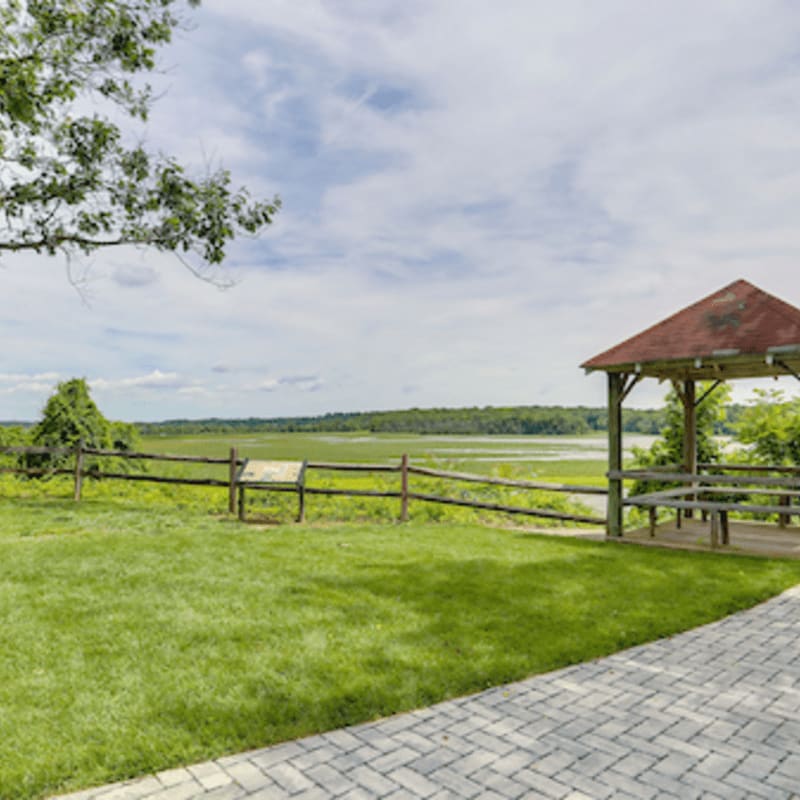 A paved walkway and green lawn lead to a wooden gazebo and fence overlooking a wide marsh under a cloudy sky.