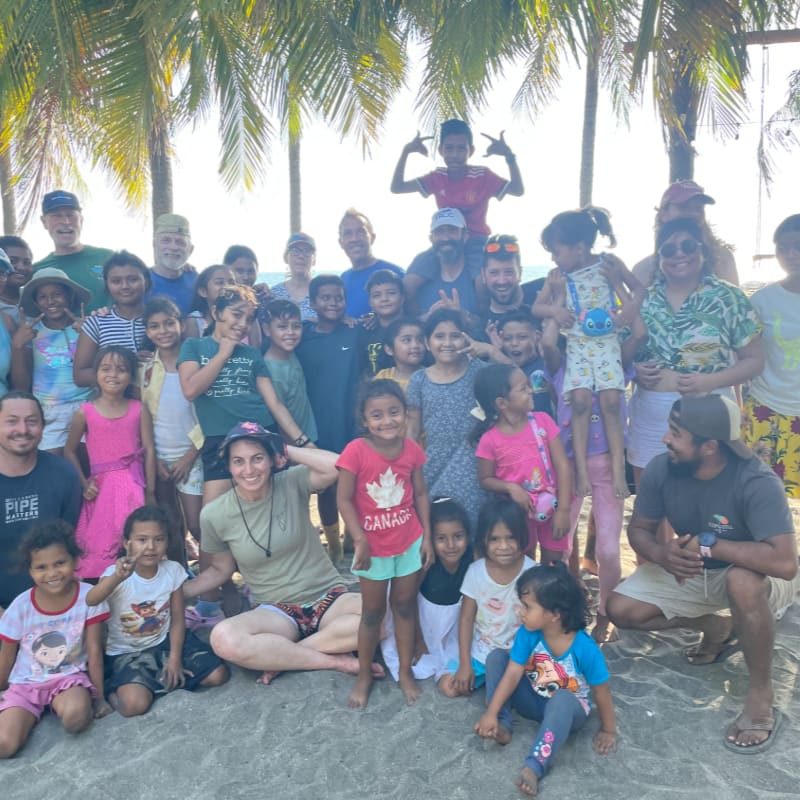 A large group of smiling children and adults pose together on a sandy beach with palm trees in the background.