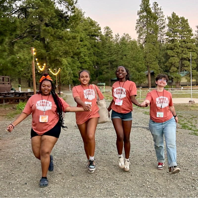 Four smiling young women in matching orange t-shirts walk hand-in-hand down a gravel path surrounded by a lush pine forest.