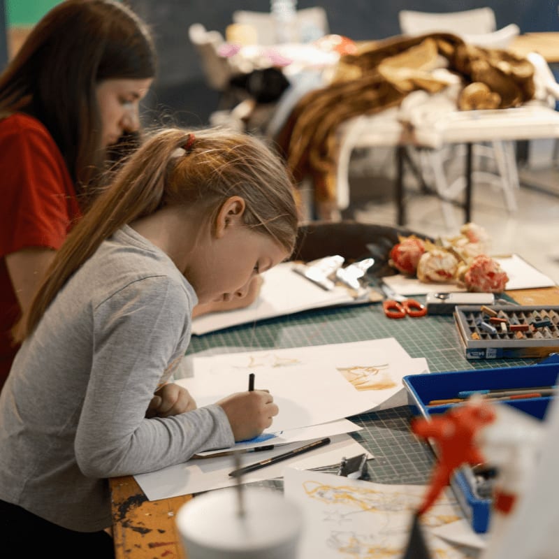A young girl draws on a piece of paper in the foreground while another girl watches and a cluttered studio fills the background.