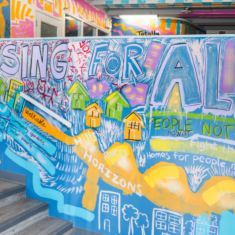 A colorful mural with text and house illustrations covers a wall in the foreground, while a brightly painted hallway extends into the background.