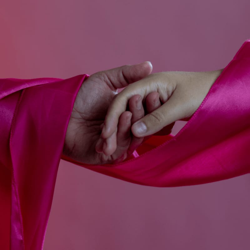 An adult hand and a child's hand hold each other, wrapped in a vibrant pink fabric against a solid purple background.