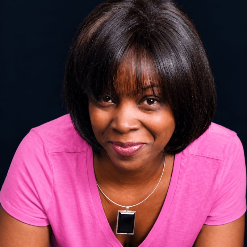 A woman wearing a pink shirt and a pendant necklace poses against a solid black background.