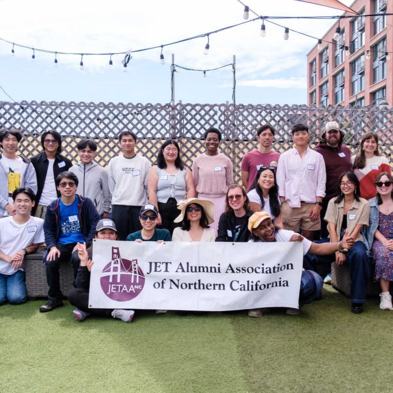 A group of people pose behind a "JET Alumni Association of Northern California" banner on a grassy area with a lattice fence and a brick building in the background.
