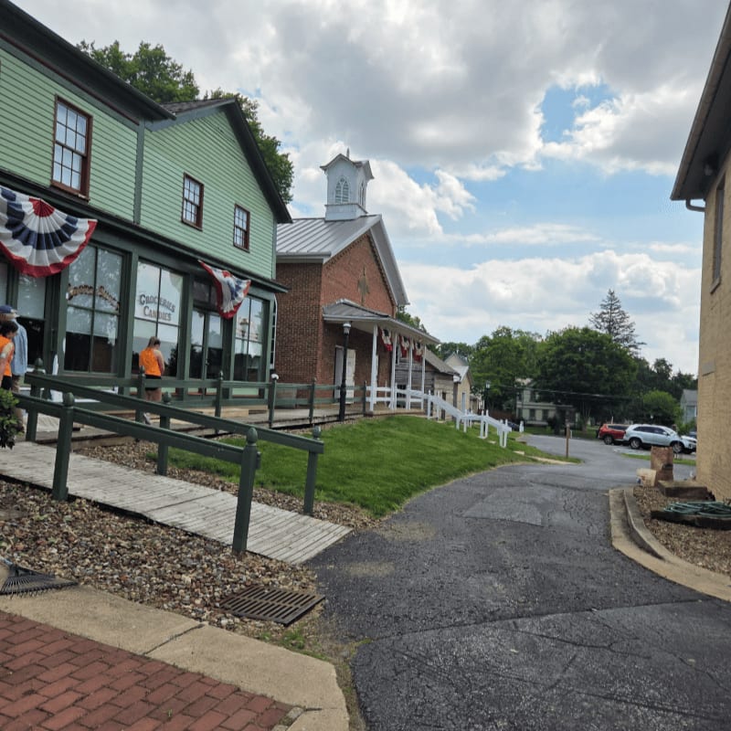 A wooden ramp leads toward a green storefront and a brick building with a cupola under a cloudy blue sky.
