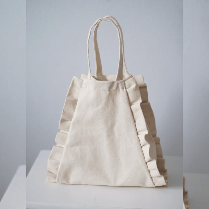 A cream-colored ruffled fabric tote bag sits on a white surface against a plain light gray background.