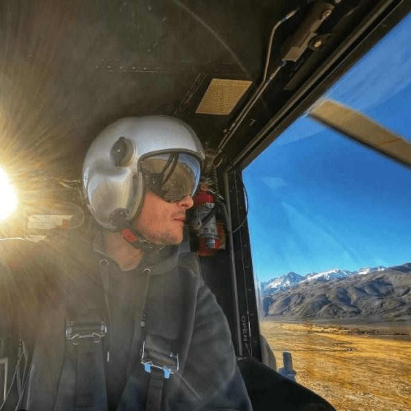 A pilot wearing a white helmet and goggles sits inside a helicopter cockpit, with a vast landscape of golden plains and snow-capped mountains visible through the window.