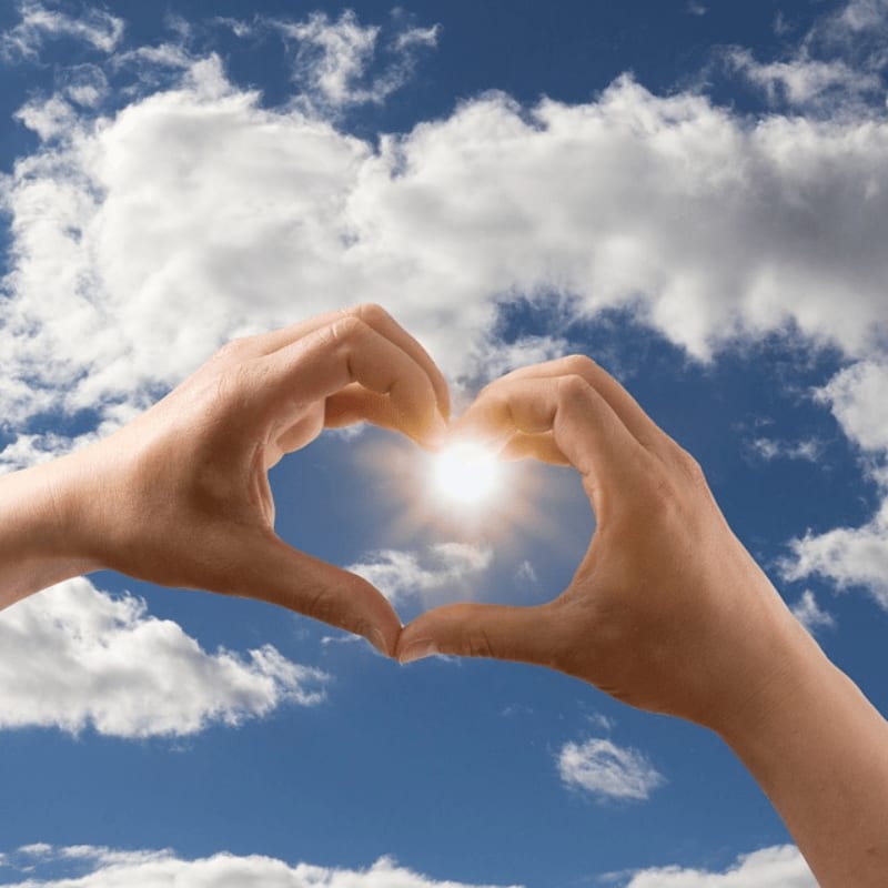 Two hands form a heart shape in the foreground, framing a bright sun against a blue sky with white clouds in the background.
