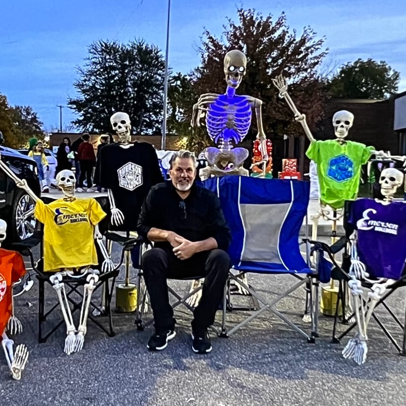 A man in black clothing sits in a chair surrounded by several skeletons dressed in colorful t-shirts in an outdoor parking lot at dusk.