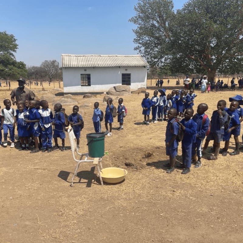 Schoolchildren in blue uniforms gather in a dirt yard with a white building and a large tree in the background.