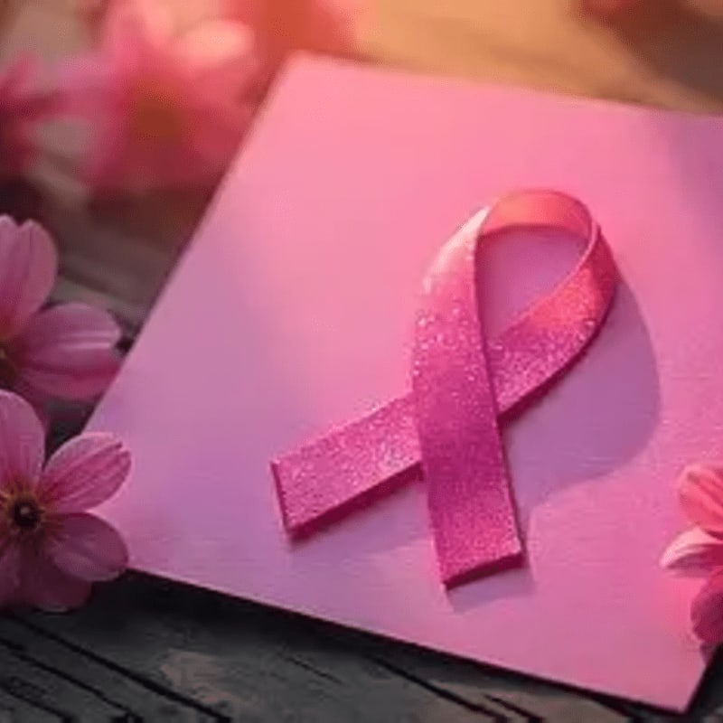 A pink awareness ribbon rests on a pink square surface, surrounded by soft pink flowers in the background.