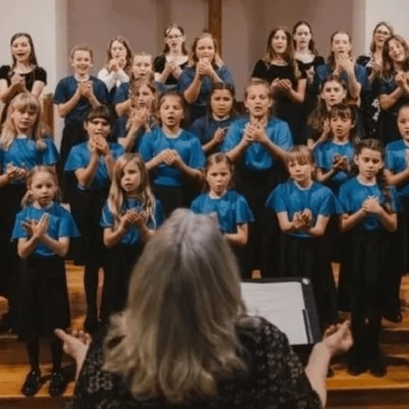 A female conductor with her back to the camera leads a choir of children dressed in blue shirts and black bottoms.