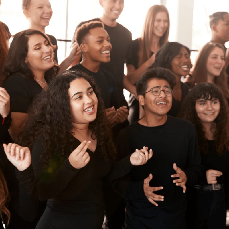A group of smiling young people in black clothing are singing and dancing in the foreground, with more cheerful friends gathered behind them in a bright room.
