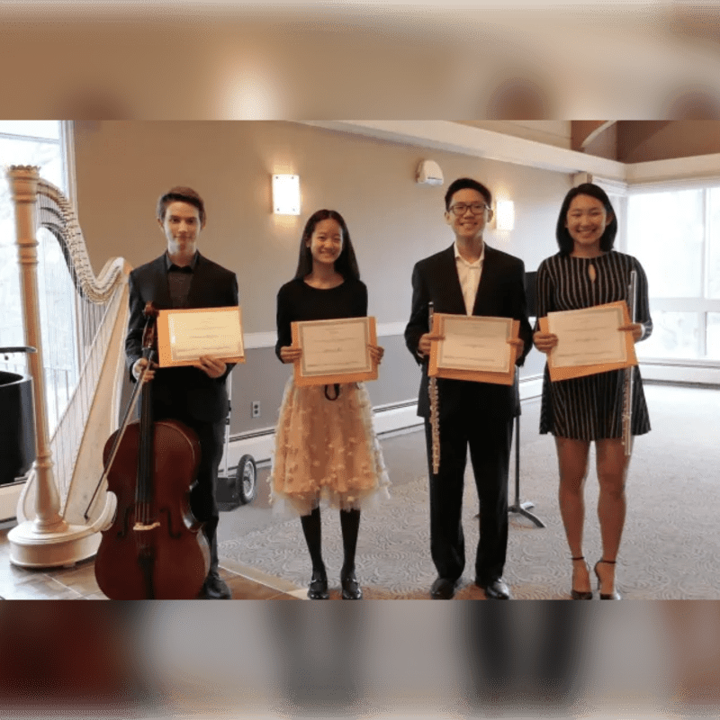 Four young people hold certificates in a room featuring a harp and a cello in the foreground against a plain wall background.