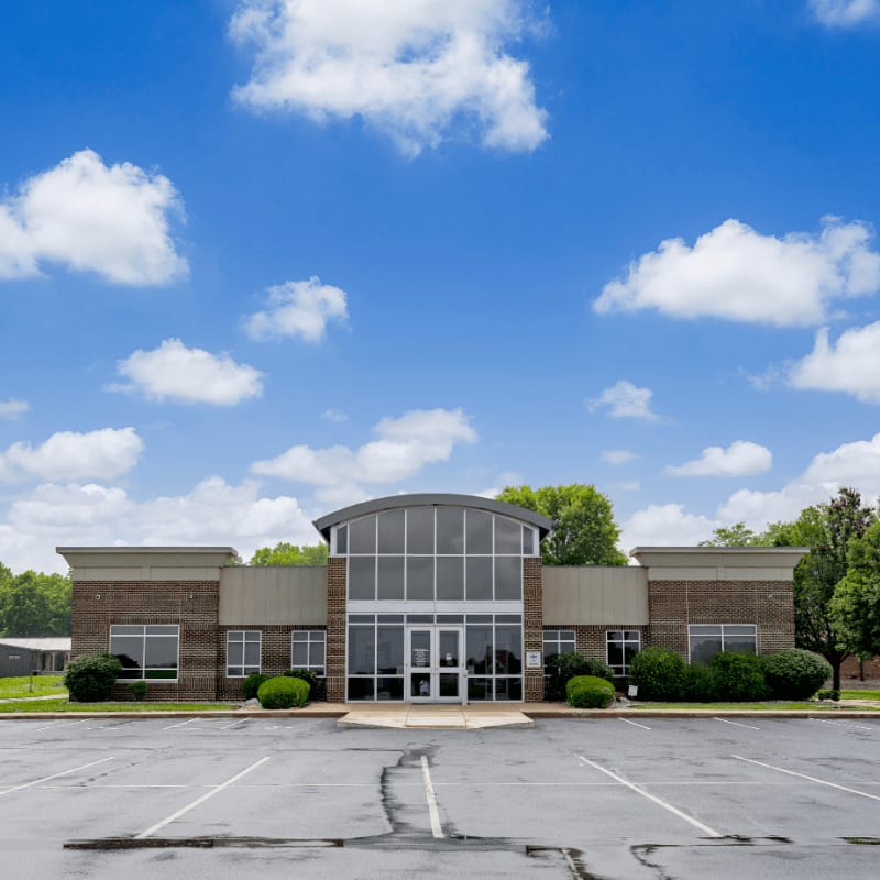 A brick office building with a curved glass entrance stands before a bright blue sky filled with fluffy white clouds and an empty asphalt parking lot in the foreground.