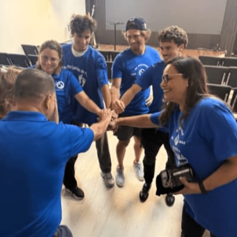 A group of people in blue shirts huddle their hands together in the foreground of a room with rows of empty chairs in the background.