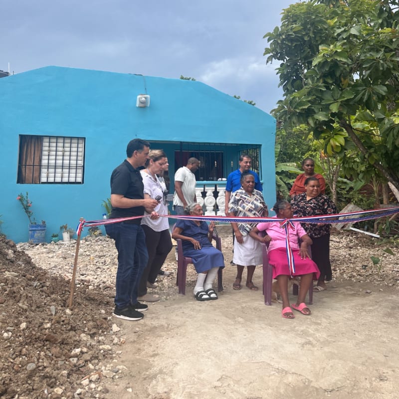 A group of people gather for a ribbon-cutting ceremony in front of a bright blue house and lush green trees.