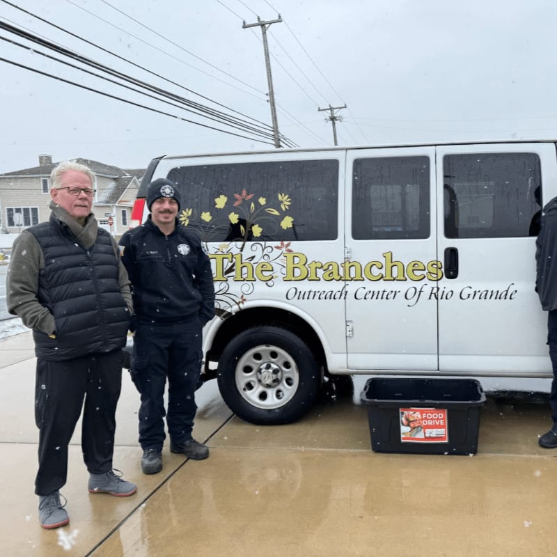 Two men stand beside a white van labeled "The Branches Outreach Center Of Rio Grande" on a wet pavement with houses and utility poles in the background.