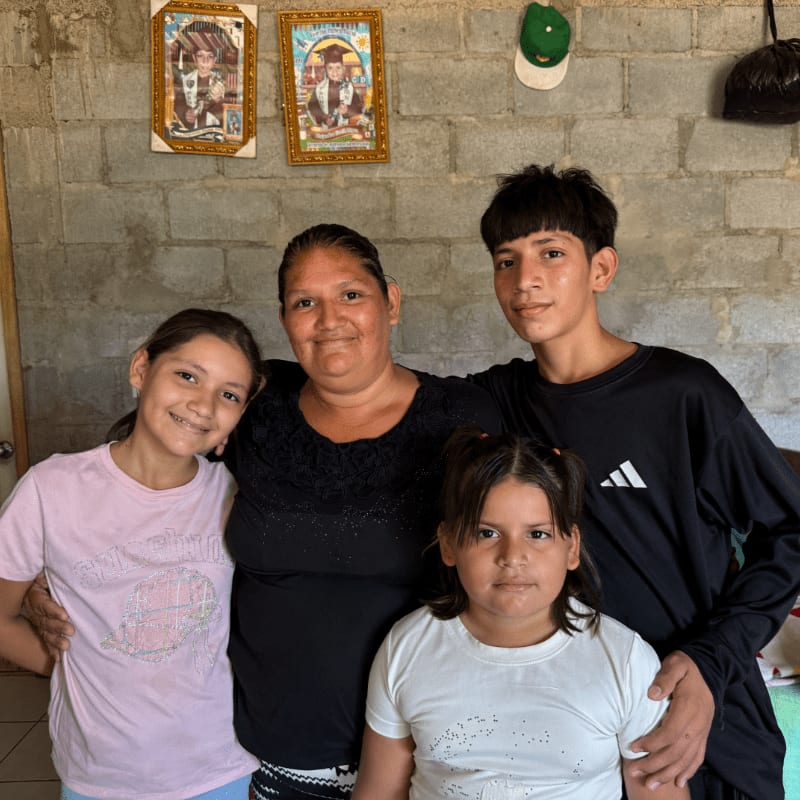 A woman and three children pose together in front of a grey cinder block wall decorated with framed pictures.