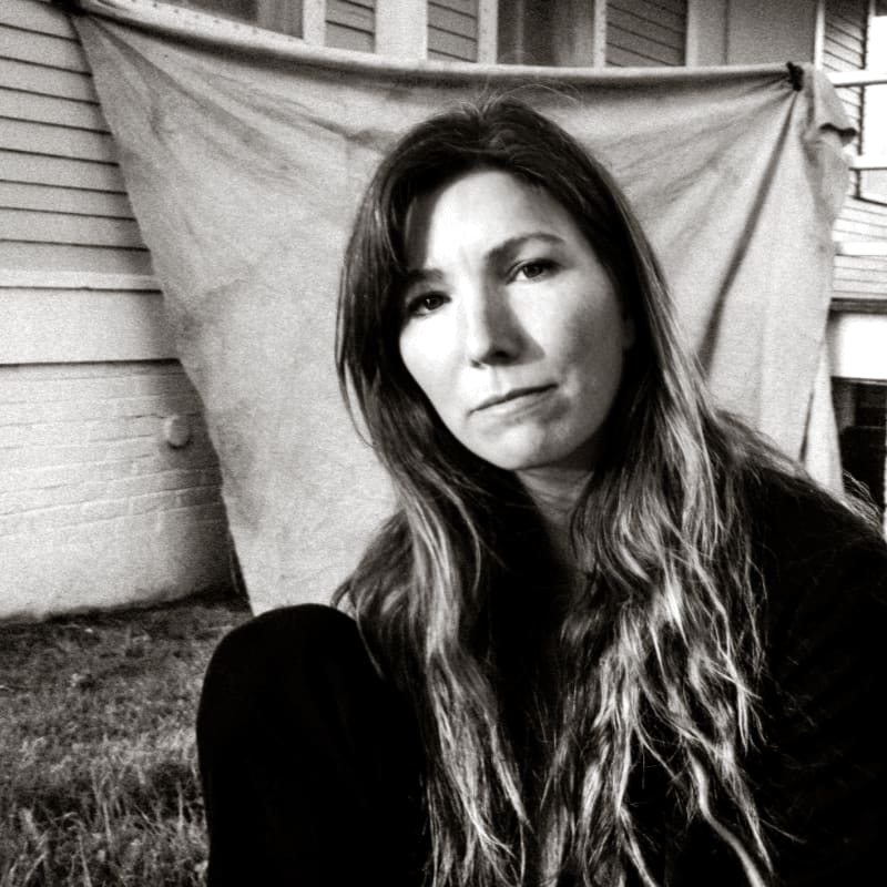 A woman with long, wavy hair sits in the foreground against a background of a draped cloth and a siding wall.