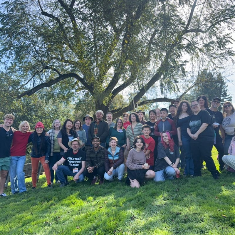 A diverse group of people pose for a photo on a grassy lawn beneath the sprawling branches of a large, leafy tree.