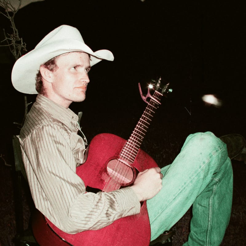 A man wearing a white cowboy hat and striped shirt plays a red guitar against a dark, nighttime background.