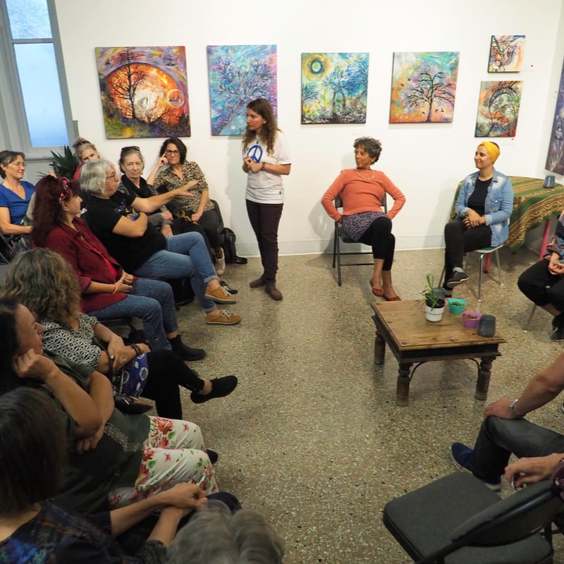 A group of women sit in a circle in an art studio, facing a woman standing in the center against a background of colorful paintings on the wall.