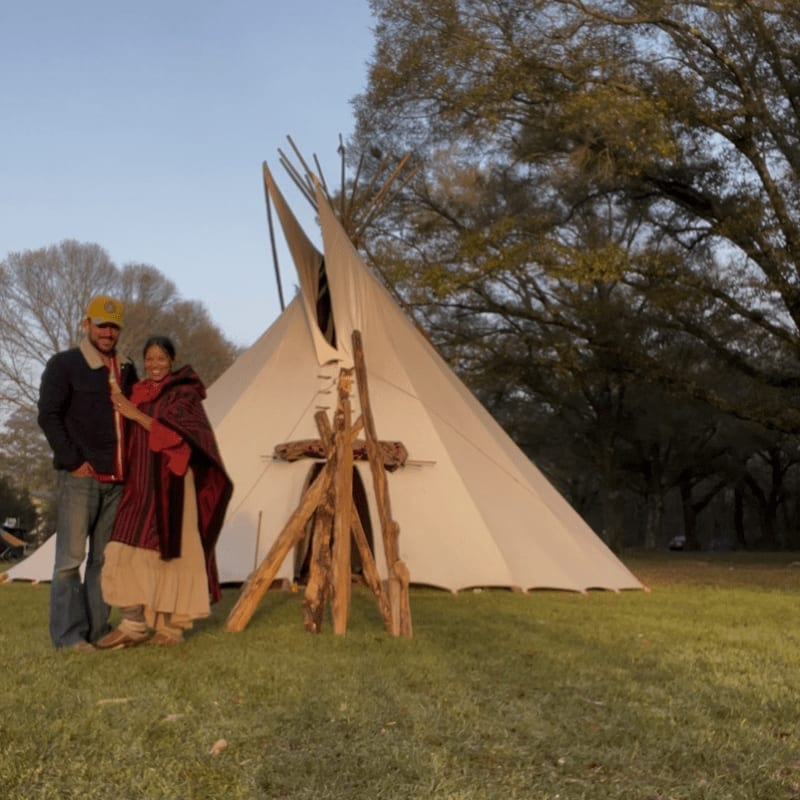 A man and woman stand beside a white tipi on a grassy field with large, leafless trees in the background.