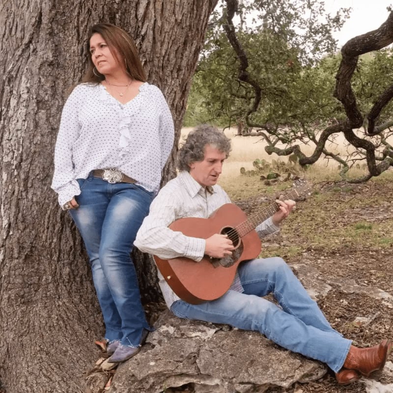 A woman leans against a large tree trunk while a man sits on a rock playing a guitar in a wooded outdoor setting.