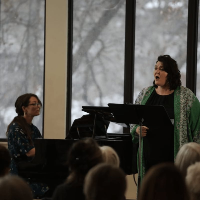 A woman sings from a music stand while another woman plays a piano in front of large windows overlooking a wintry landscape.