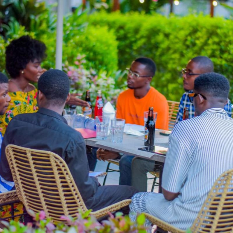 A group of people sit around a table with drinks in the foreground, set against a lush background of green hedges.