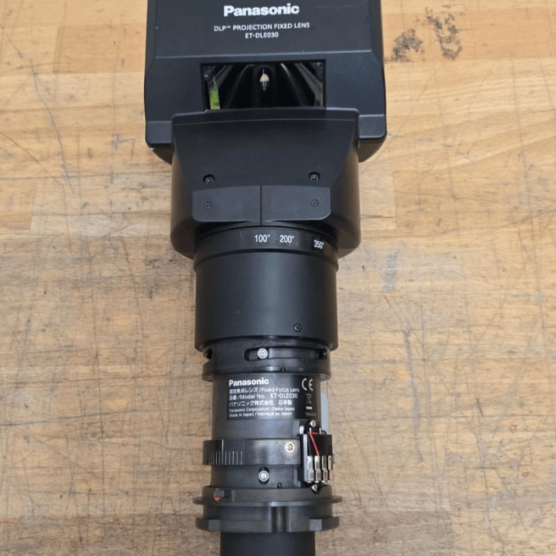 A black Panasonic projection lens sits vertically on a light-colored wooden surface.