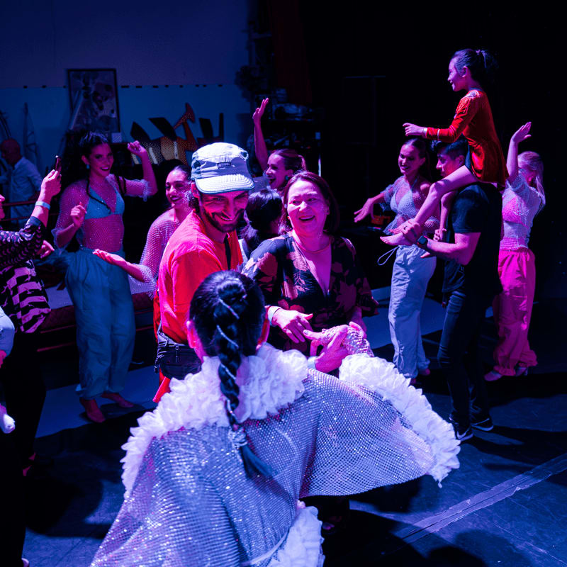 A group of people dance and socialize in a dimly lit room with a dark background.