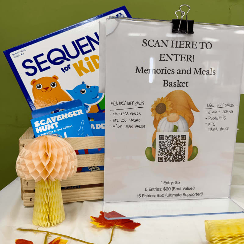 A sign for a "Memories and Meals Basket" raffle stands in the foreground next to a honeycomb mushroom decoration, with a board game and scavenger hunt kits in a wooden crate in the background.