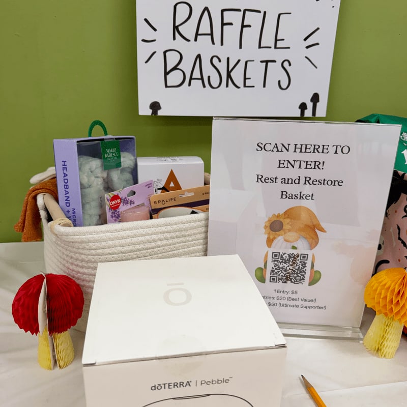 A white raffle basket filled with wellness products and a QR code sign sit in the foreground against a green wall with a "RAFFLE BASKETS" sign in the background.