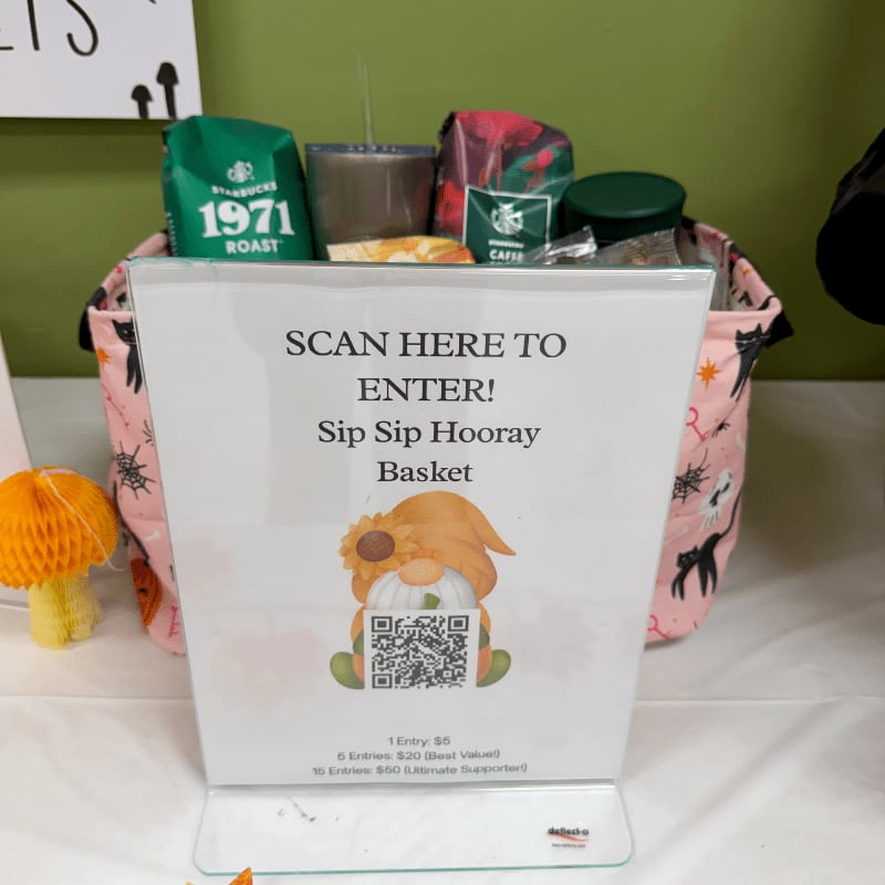 A sign for a "Sip Sip Hooray Basket" raffle stands in the foreground, with a gift basket filled with coffee products and a small orange mushroom decoration set against a green wall.