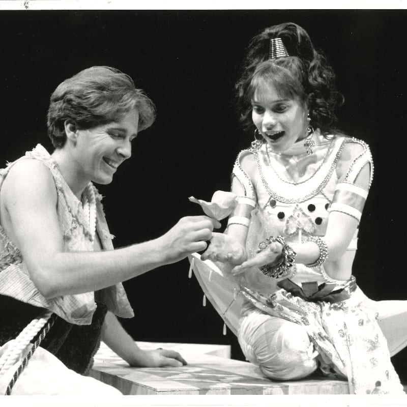 A smiling man in a simple tunic offers a flower to a woman in an ornate, sparkling costume against a dark background.