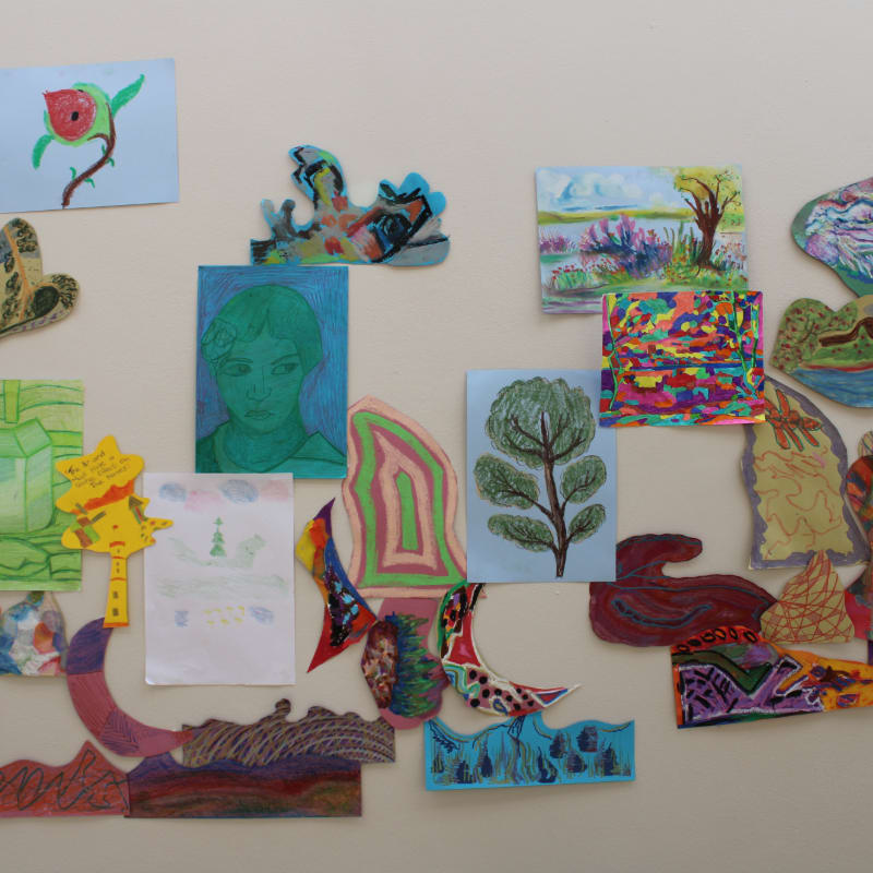 Various colorful children's drawings and cut-out art pieces are arranged across a plain, light-colored wall.
