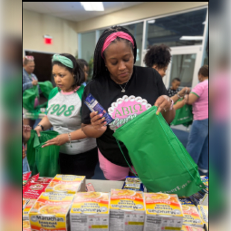 A woman in a black t-shirt fills a green bag with food items in the foreground, while other volunteers and people stand in a brightly lit indoor room in the background.
