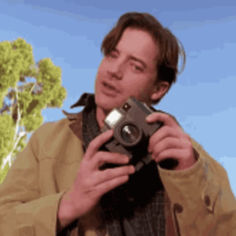 A young man in a tan jacket holds a camera in the foreground, with a bright blue sky and a green tree in the background.