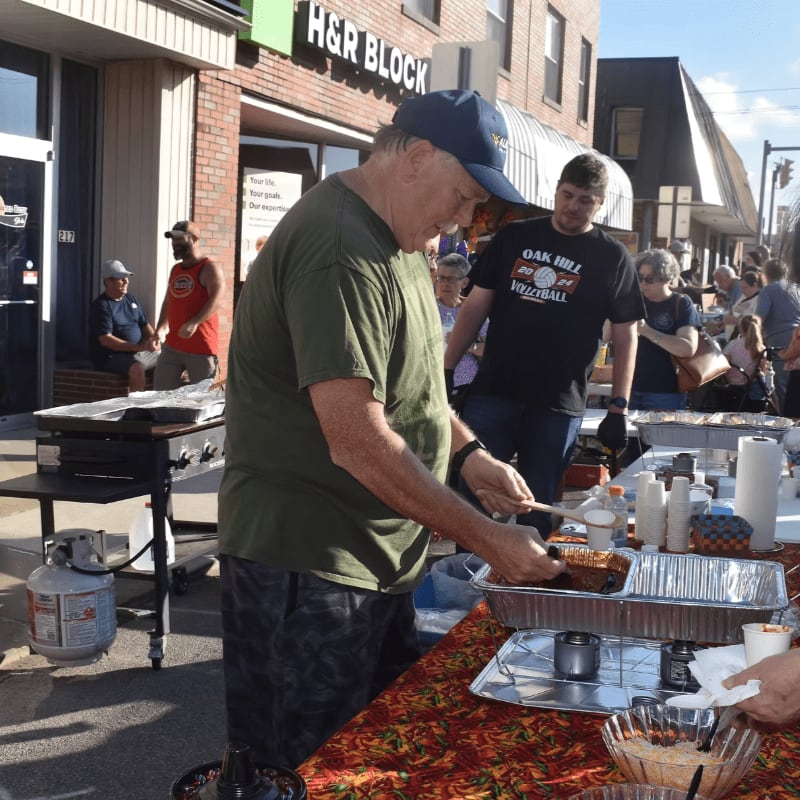 A man in a green shirt and blue cap prepares food at an outdoor table in the foreground, with other people and an H&R Block storefront in the background.