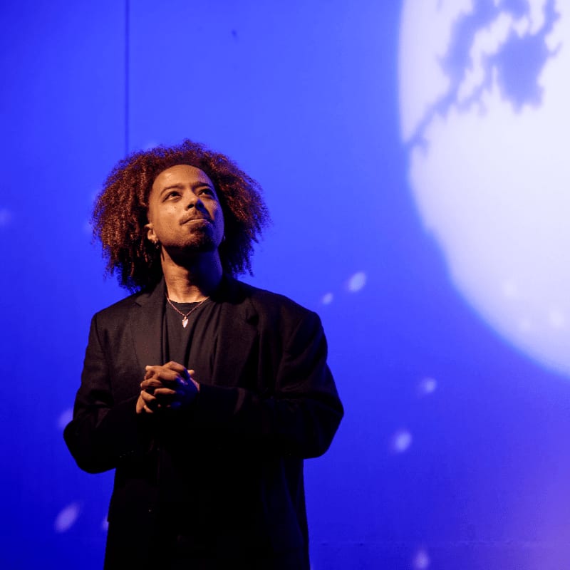 A man with curly hair and a black blazer stands with clasped hands against a deep blue background featuring a large, bright circular light.