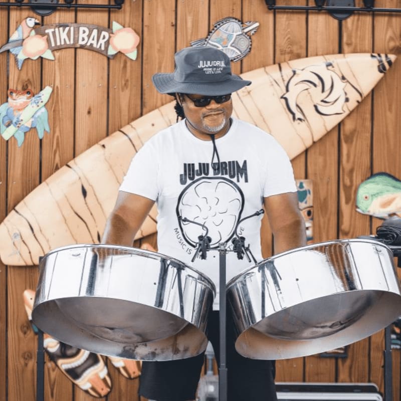 A man in a hat and t-shirt plays steel drums in front of a wooden wall decorated with a surfboard and tiki signs.