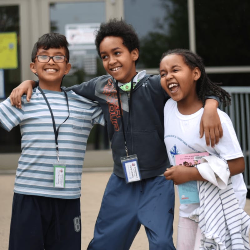 Three smiling children stand arm-in-arm in the foreground in front of a blurred building entrance.