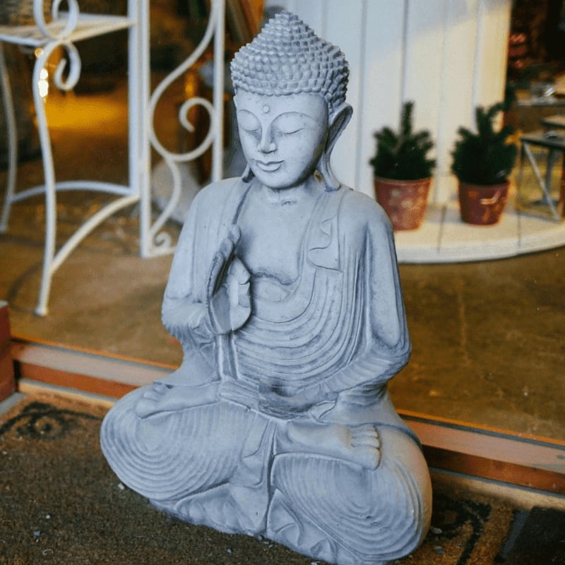 A grey Buddha statue sits in the foreground with a white metal stand and potted plants in the blurred background.